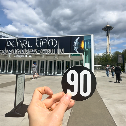 90 The Original sticker held in front of the Space Needle and Climate Pledge Arena in Seattle, featuring Pearl Jam concert information.