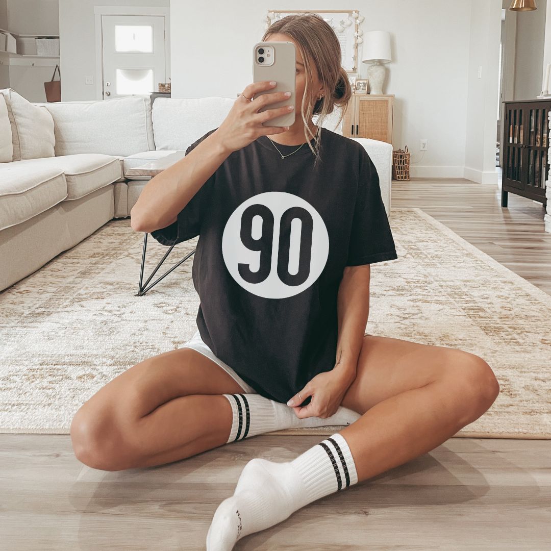 Casual chic at home, woman in a black 90 The Original logo tee taking a selfie, embodying relaxed modern style.