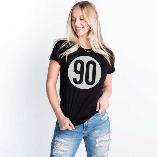 Smiling woman in a black 90 The Original tee and distressed jeans, showcasing everyday style with a '90s twist.